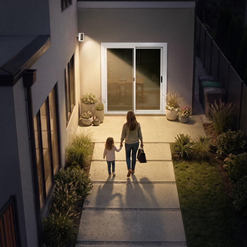Mother and child walking toward a lit patio entrance at night, illuminated by a wall-mounted spotlight for safe arrival